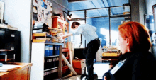 a man is standing in a room with a stack of folders on a shelf and a woman is sitting in a chair