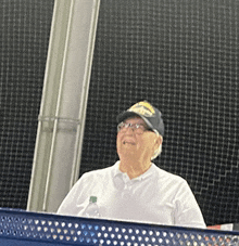 an elderly man wearing a hat and glasses stands behind a fence