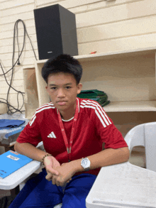 a boy wearing a red adidas shirt sits in a classroom