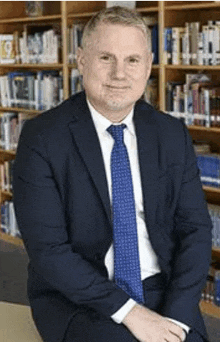 the man is wearing a suit and tie and is sitting in front of a bookshelf .