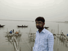 a man wearing glasses stands in front of a body of water with boats in the background