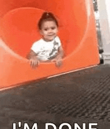 a little girl is sitting on top of an orange slide at a playground .