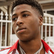 a young man wearing a red shirt and a silver chain around his neck is standing in front of a white fence .