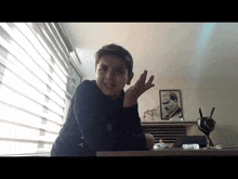 a young boy is sitting at a desk in front of a window with his hand up .