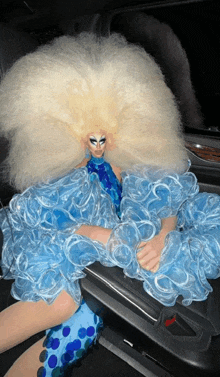 a drag queen with a very large blonde wig sits in a car