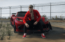 a man in a red jacket is sitting on the hood of a red sports car