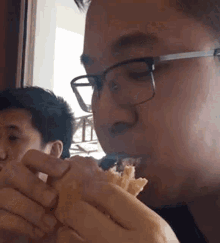 a man wearing glasses is eating a sandwich while another man watches .