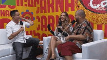 two men and a woman are sitting on a couch with microphones in front of a wall that has the word carn on it