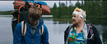 a man in a blue jacket and a woman in a colorful shirt are standing next to each other in front of a lake