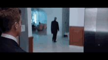 a man in a suit is standing in a hallway looking at a man in a suit walking down the hallway