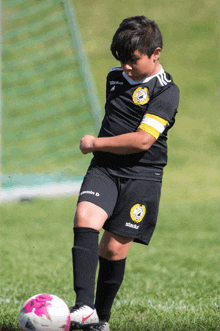 a young boy wearing a black shirt with a yellow emblem on the front kicking a soccer ball