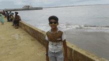 a boy wearing sunglasses stands on a stone wall near the water