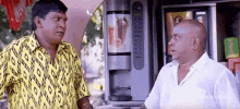 two men are standing next to each other in front of a vending machine and talking to each other .