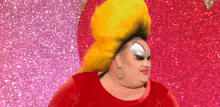 a drag queen with a big yellow wig and earrings is standing in front of a pink and purple background .