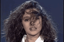 a woman with curly hair is wearing a white shirt and a black jacket and has her hair blowing in the wind .