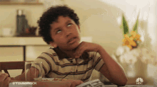 a young boy is sitting at a table with a newspaper and a glass of water with the nbc logo in the background