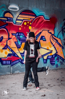 a woman wearing a black t-shirt with the letter a on it stands in front of a wall with graffiti on it