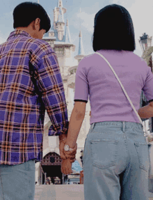 a man in a plaid shirt and a woman in a purple shirt hold hands in front of a castle