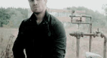 a man in a black jacket is standing in a field next to a water pump .