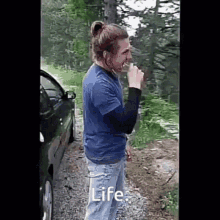 a man in a blue shirt is smoking a cigarette while standing next to a car on a dirt road .