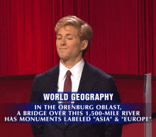 a man in a suit and tie stands in front of a red curtain and a sign that says world geography