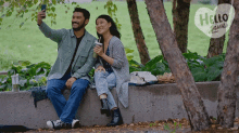 a man and a woman are sitting on a wall and taking a selfie with a hello again sign behind them