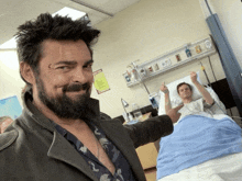 a man with a beard is standing next to a man in a hospital bed giving the middle finger