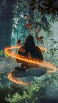 a man in a hooded sweatshirt sits in a lotus position surrounded by flames and playing cards