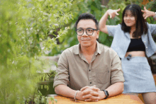 a man wearing glasses sits at a table with his hands folded while a woman in a blue shirt stands behind him