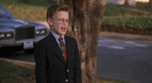 a young boy wearing a suit and tie is standing in front of a tree .