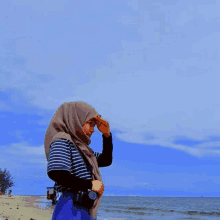 a woman wearing a hijab and a striped shirt stands on a beach holding a camera