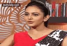 a woman in a red top and black and white saree is standing in front of a book shelf .