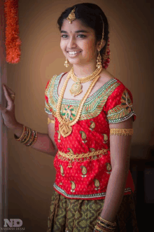 a woman wearing a red top and gold jewelry is standing in front of a wall with the letters nd on the bottom right