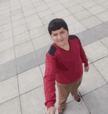 a young man wearing a red sweater and brown pants stands on a tiled sidewalk