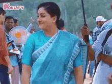 a woman in a blue saree is smiling and holding an umbrella in front of a crowd of people .