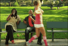 a woman sitting on a park bench drinking a bottle of water while a girl in pink shorts runs past her