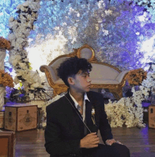 a man in a suit sits in front of a couch with flowers