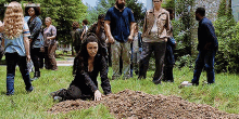 a woman is kneeling in the grass next to a pile of ants while a group of people are standing around her .