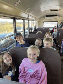 a girl wearing a pink barbie shirt sits on a school bus