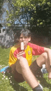 a young man in a yellow and red shirt is eating an ice cream bar while sitting in the grass .