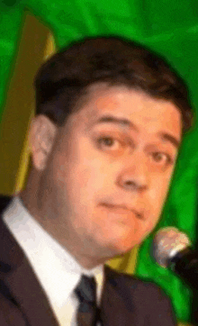 a man in a suit and tie is standing in front of a microphone with a green background .