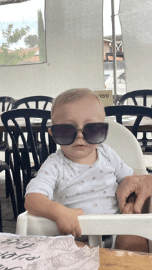 a baby wearing sunglasses sits in a high chair in front of a sign that says ' hebrew ' on it