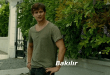 a man in a green t-shirt is standing in front of a bush with the word bakilir written below him