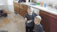 two young boys are standing next to each other in a kitchen with a lot of food on the floor .