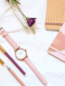 a pink watch sits next to a pen a notebook and flowers