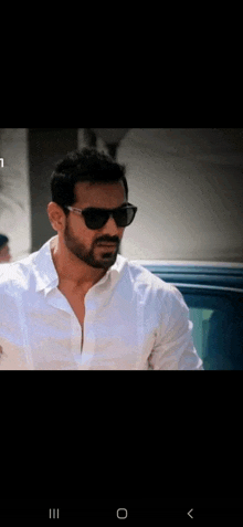 a man wearing sunglasses and a white shirt stands in front of a car .