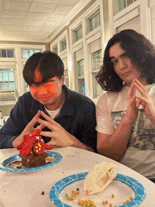 two young men are sitting at a table with plates of food and one has a cake on his face