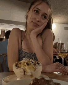 a woman sits at a table with her hand on her chin and a bowl of food in front of her