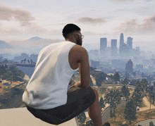 a man in a white tank top sits on the edge of a building looking out over a city