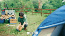 a man in a green shirt is kneeling in front of a blue tent with the words get real written on the bottom